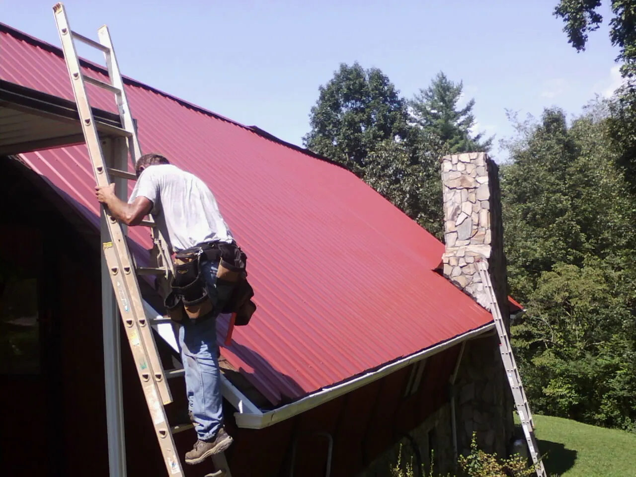 Roofer on a ladder beside a red metal roof and stone chimney — Soffit & Fascia Repair by Roofing Brothers in Clarkson